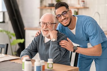 Elderly man and healthcare worker smiling together at a table with medicine bottles.
