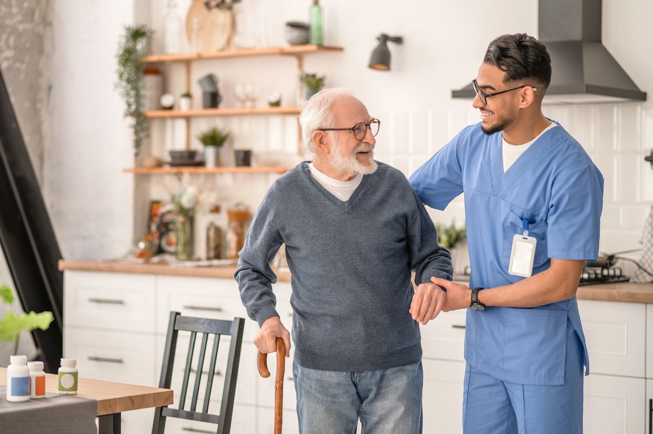 A caregiver helps an elderly man walk with a cane in a cozy kitchen.