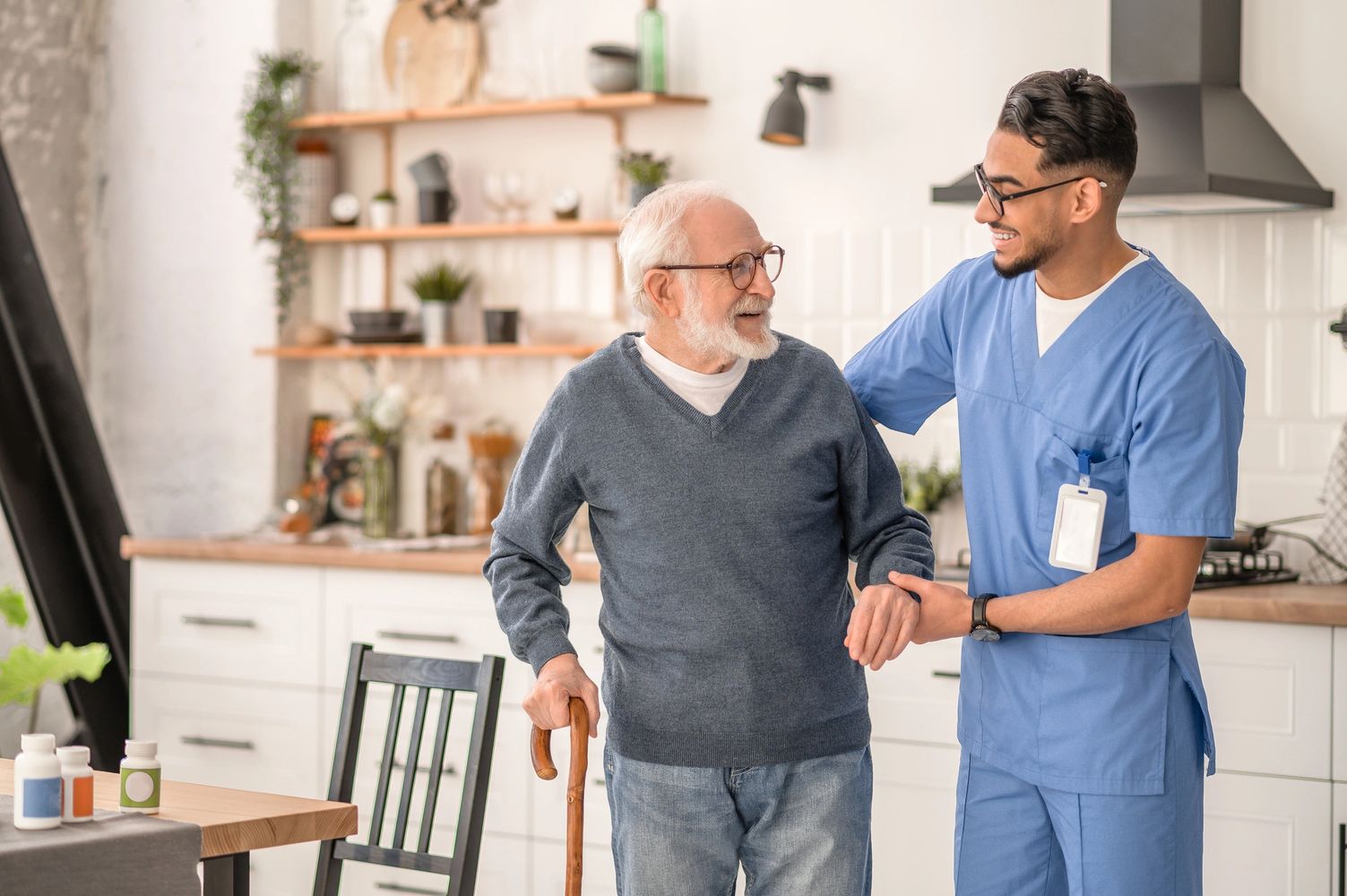 A young nurse assists an elderly man with a cane in a cozy kitchen - MedGoPlus