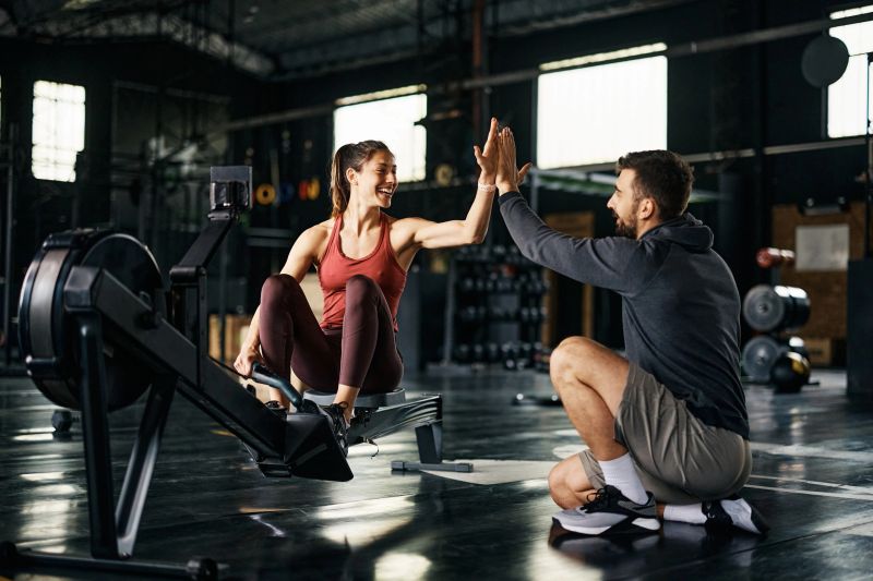 Supportive fitness instructor giving high-five to athletic woman after exercising on rowing machine during cross training in a gym.