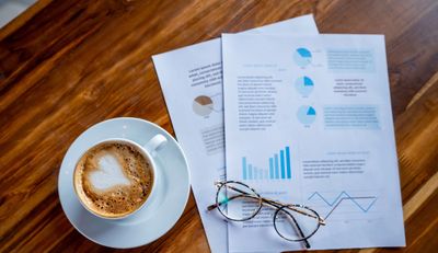 A cup of coffee, eyeglasses, and printed charts on a wooden table.