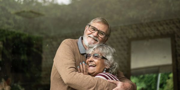 Elderly couple smiling and hugging warmly indoors near a window.