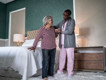 A nurse helps an elderly woman stand up beside a bed.