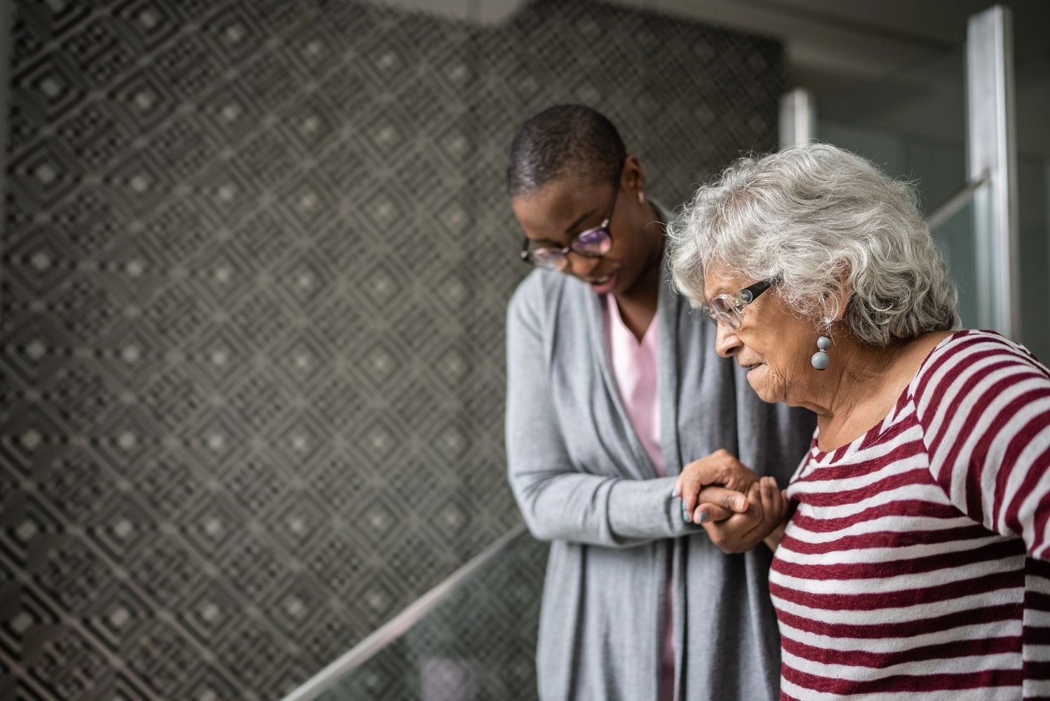 A caregiver assists an elderly woman walking indoors.