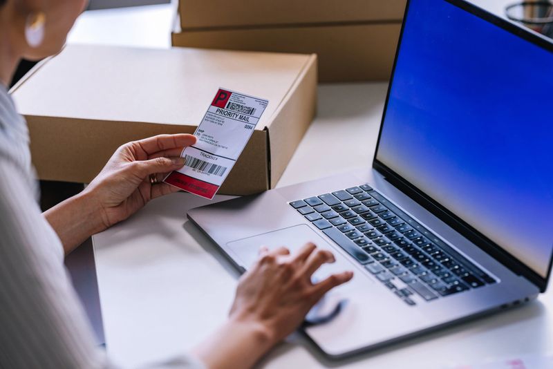 A side view of an anonymous female Caucasian entrepreneur holding a label and checking customer's details on the computer before shipping the merchandise in a cardboard box.