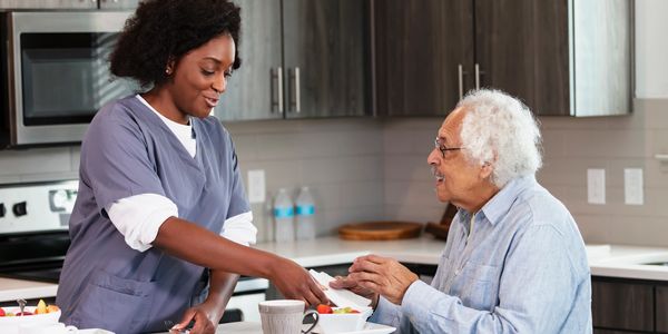 Caregiver hands food to elderly man in kitchen.