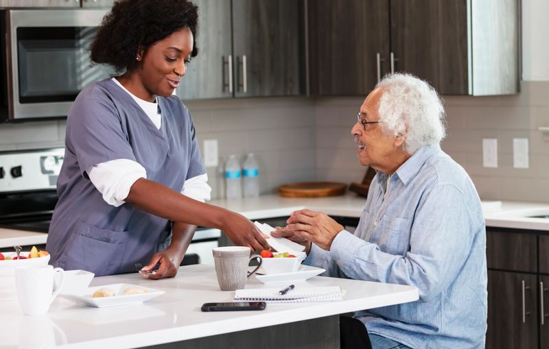 A home healthcare worker, an African-American woman in her 30s, helping a senior man in his 80s, have a healthy snack in the kitchen. He is Hispanic and Caucasian.