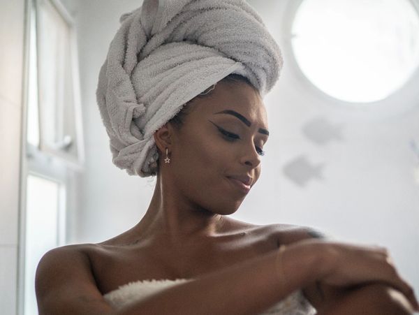 Woman with towel turban applying lotion in bathroom.