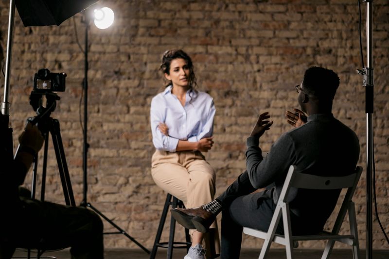 Unrecognizable male journalist interviewing young attractive woman sitting on a bar stool with her arms crossed and listens to the question