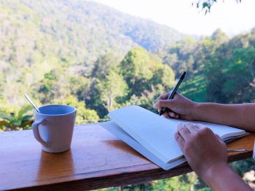 Person writing in a notebook outdoors with a cup nearby.