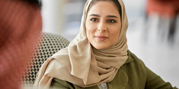 A woman wearing a beige hijab and green coat attentively listening.