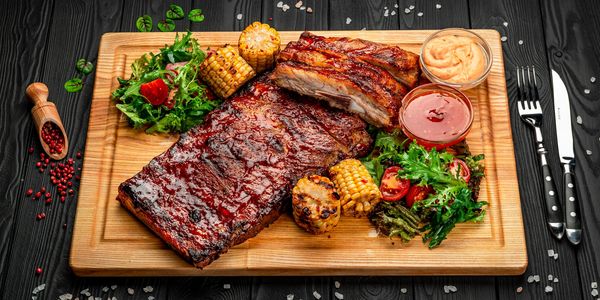 Grilled barbecue ribs with corn, salad, and sauces on a wooden board.