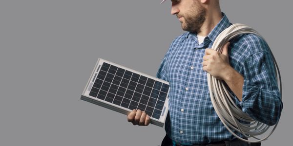 Solar technician holding a solar panel and coiled wire, wearing a hard hat and tool belt.