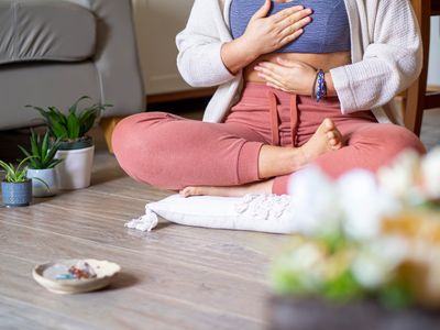 Person meditating indoors with hands on chest and abdomen, sitting cross-legged on a cushion.