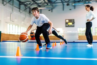 Child dribbling basketball through cones indoors.