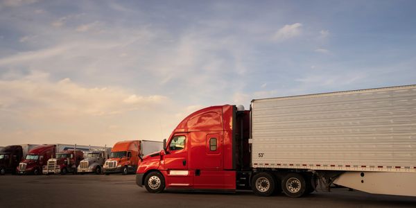 A bright red semi-truck with a trailer parked among other trucks at sunset.