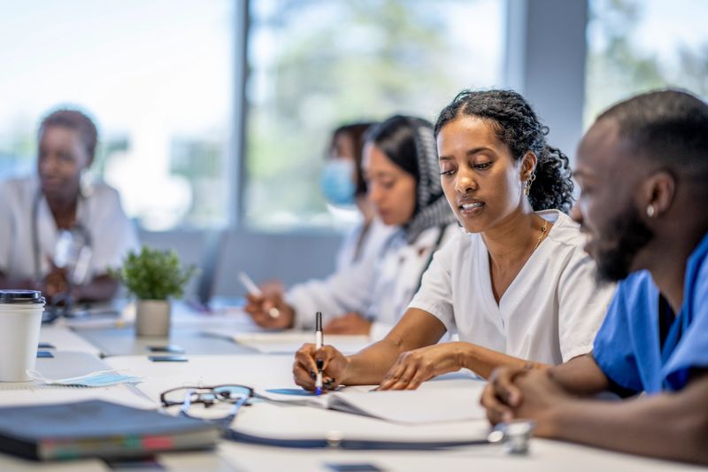 A small group of medical professionals sit around a boardroom table as they meet to discuss patient cases.  They are each dressed professionally in scrubs and lab coats as they work through the case files spread out on the desk collaboratively in small groups.