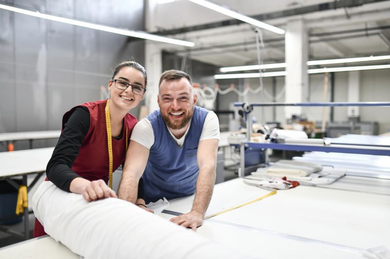 Portrait Of Male And Female Textile Factory Colleagues Measuring And Spreading Material Rolls