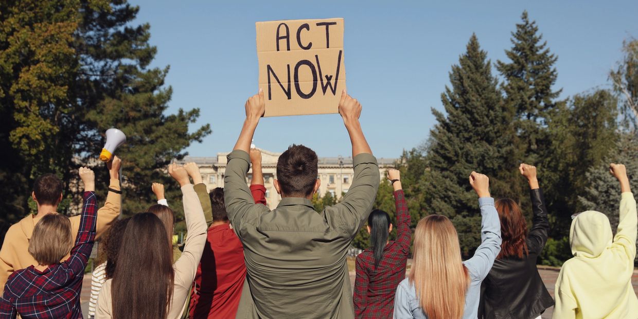 Group of people raising fists with one holding an "ACT NOW" sign outdoors.