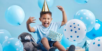 Joyful boy wearing a party hat surrounded by blue balloons and presents.