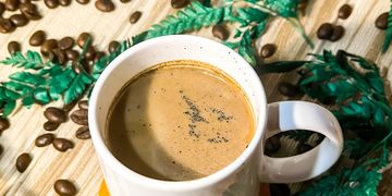 A white cup of coffee surrounded by coffee beans and green leaves.