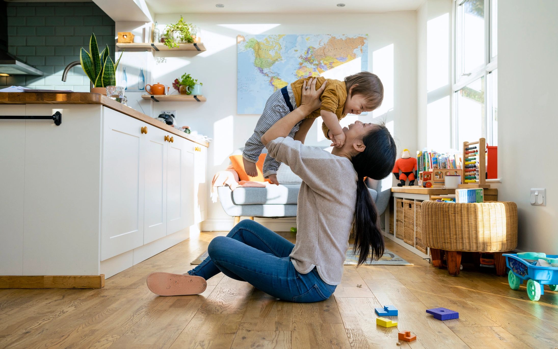 A mother joyfully lifts her child in a sunlit living room filled with toys and plants.