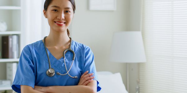 Smiling nurse in blue scrubs with stethoscope, standing confidently in a bright medical room.