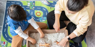 Two children playing with sand and toys in a box on a colorful mat.