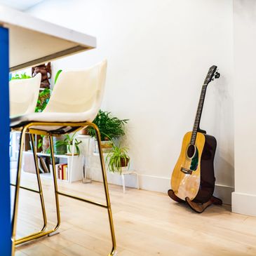 Acoustic guitar resting on a stand in a bright, modern room with plants and chairs.