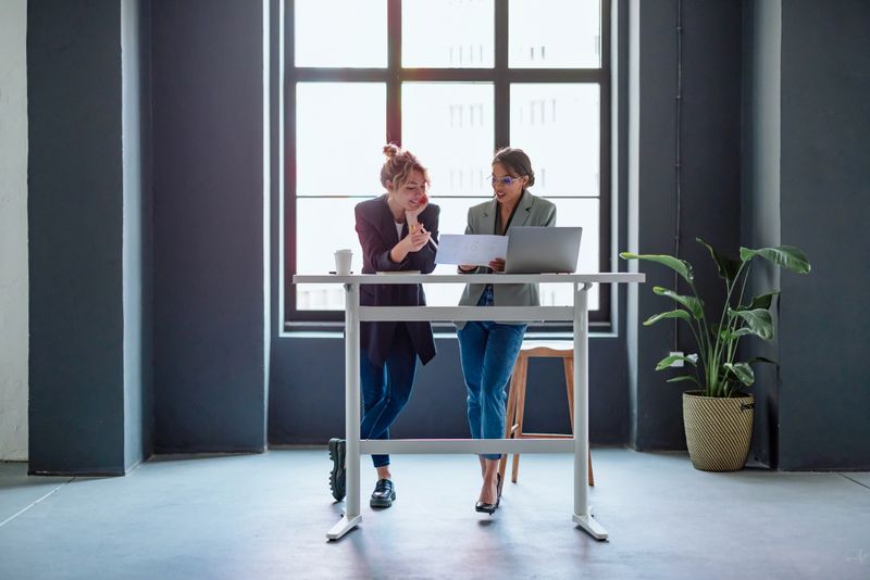 Smiling females colleagues using laptop computer, holding documents and talking about business strategy while standing together at office desk.
