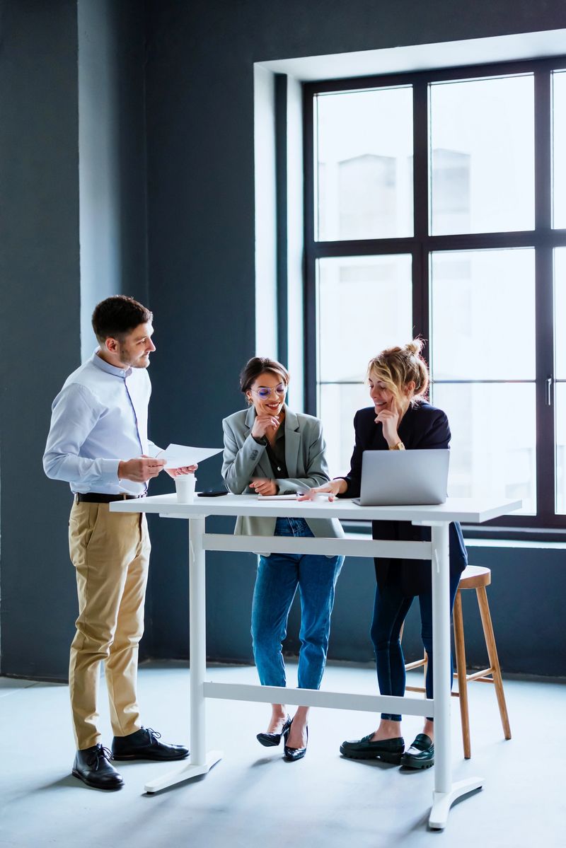 Team of three colleagues using laptop computer and business papers while discussing ideas and laughing during a casual meeting in the modern office with big window.