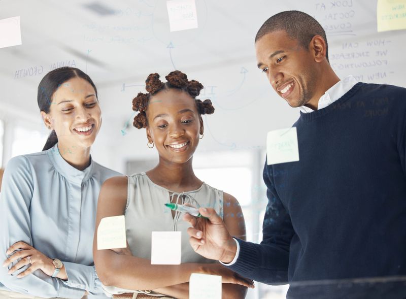 Three happy diverse businesspeople writing ideas and brainstorming together in an office at work. Cheerful hispanic businessman making a note on a glass window with female coworkers