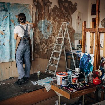 Artist painting on a large canvas in a cluttered studio.
