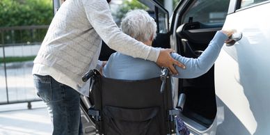 Caregiver helping elderly woman in wheelchair into a car.