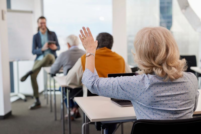 A senior woman is asking for an explanation in the computer course while sitting with elderly students in the classroom.
