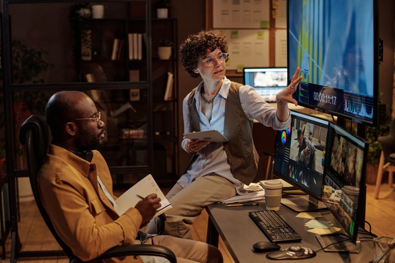 Young project manager pointing at big screen and explaining the idea of shooting to designer while he writing in notebook