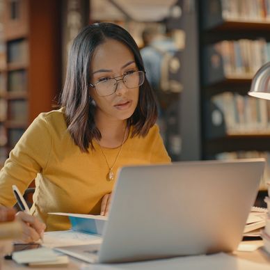 A woman studies intently with a laptop and books in a library.