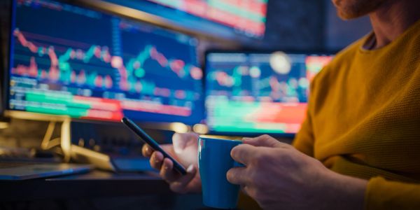 Person holding a coffee mug and smartphone with stock market charts on screens.
