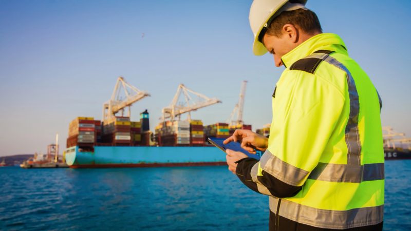 Foreperson in protective gear using digital tablet in front of container terminal port