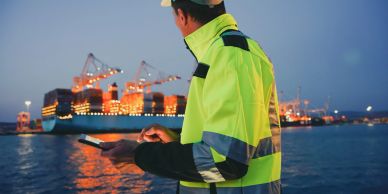 Worker in safety gear using a tablet near a lit cruise ship at night.