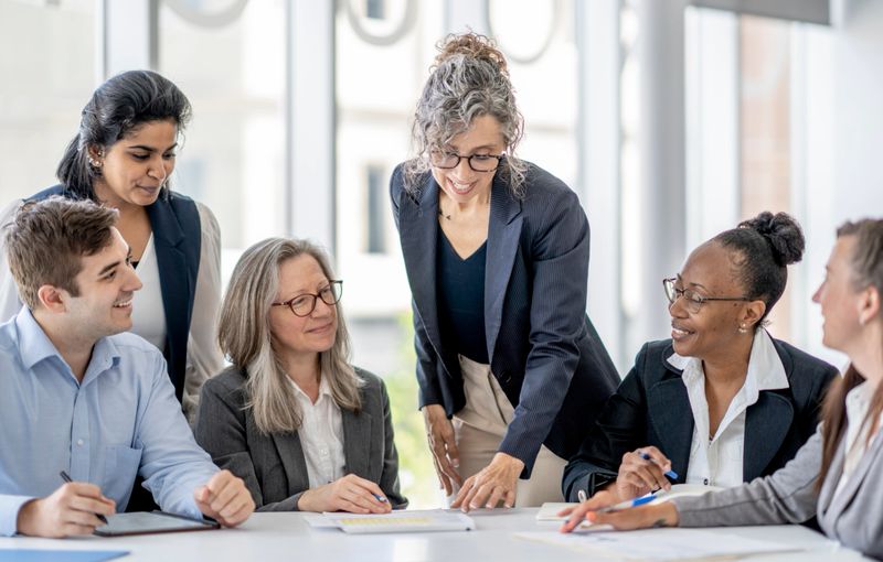 A small group of business professionals sit around a table as they meet to discuss the future of the company.  They are all dressed in business attire and have various papers and files spread out on the table in front of them as they refer to them during the meeting.
