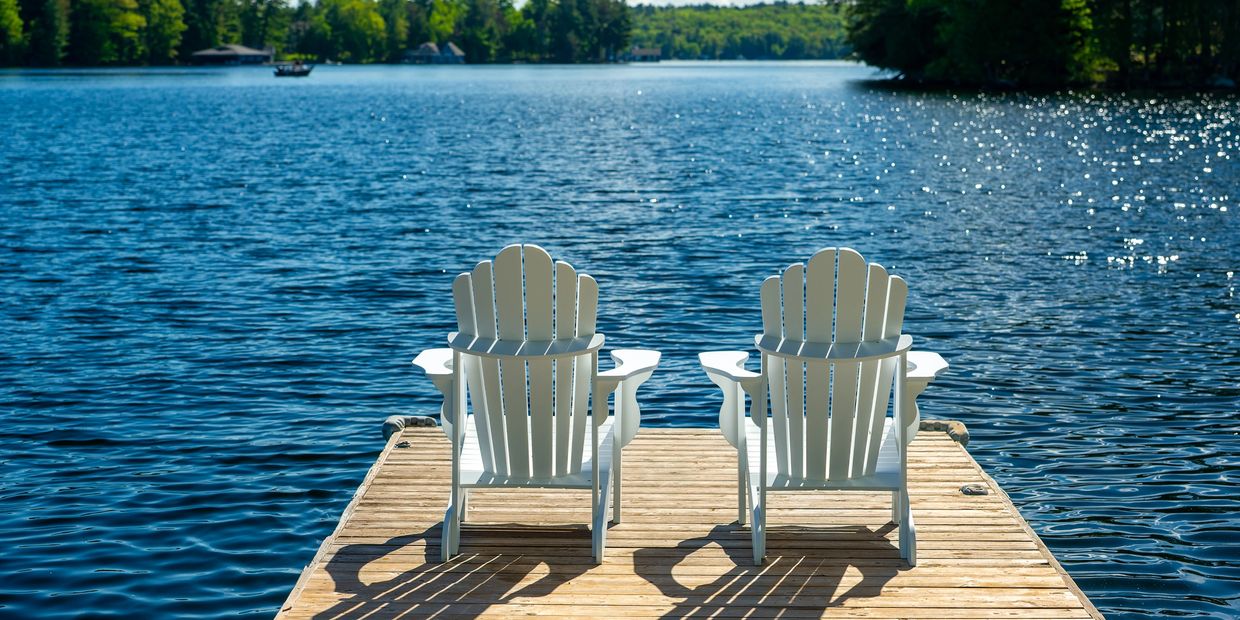 Two white Adirondack chairs on a wooden dock overlooking a peaceful lake.