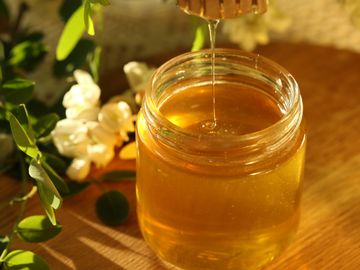 Golden honey dripping from a wooden dipper into a glass jar surrounded by flowers.