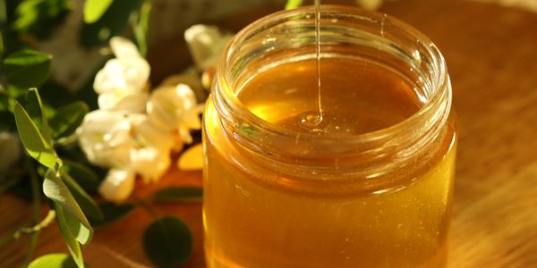 Golden honey dripping from a wooden dipper into a glass jar surrounded by flowers.