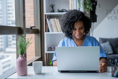 A woman in blue scrubs working on a laptop at a bright desk.