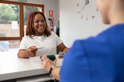 A smiling woman hands a card to a receptionist in a blue uniform.