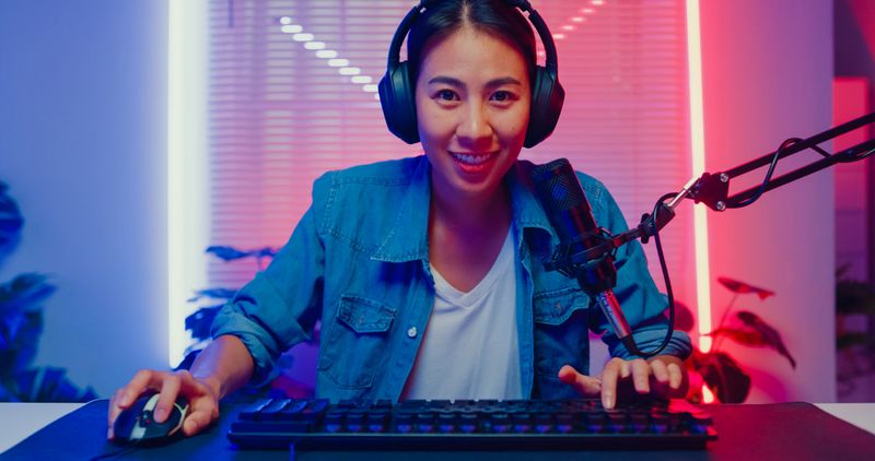 Young Asian woman playing online computer video game and broadcast streaming live in neon lights living room at home. Gamer lifestyle concept.