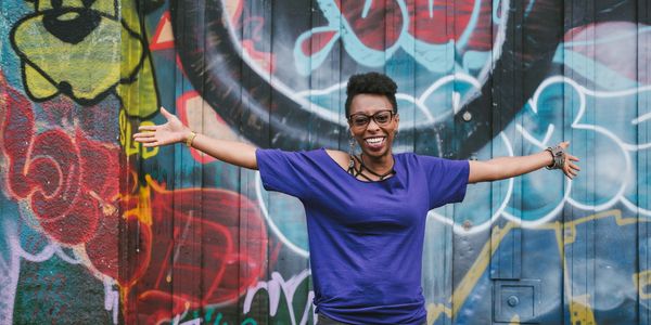 Smiling person in blue shirt with arms outstretched against graffiti wall.