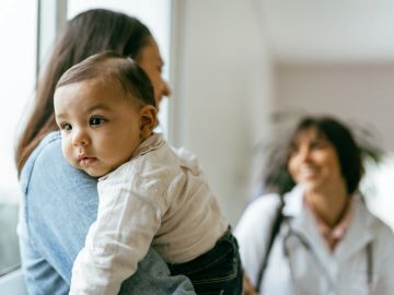 A mother holds her baby while a doctor smiles in the background.