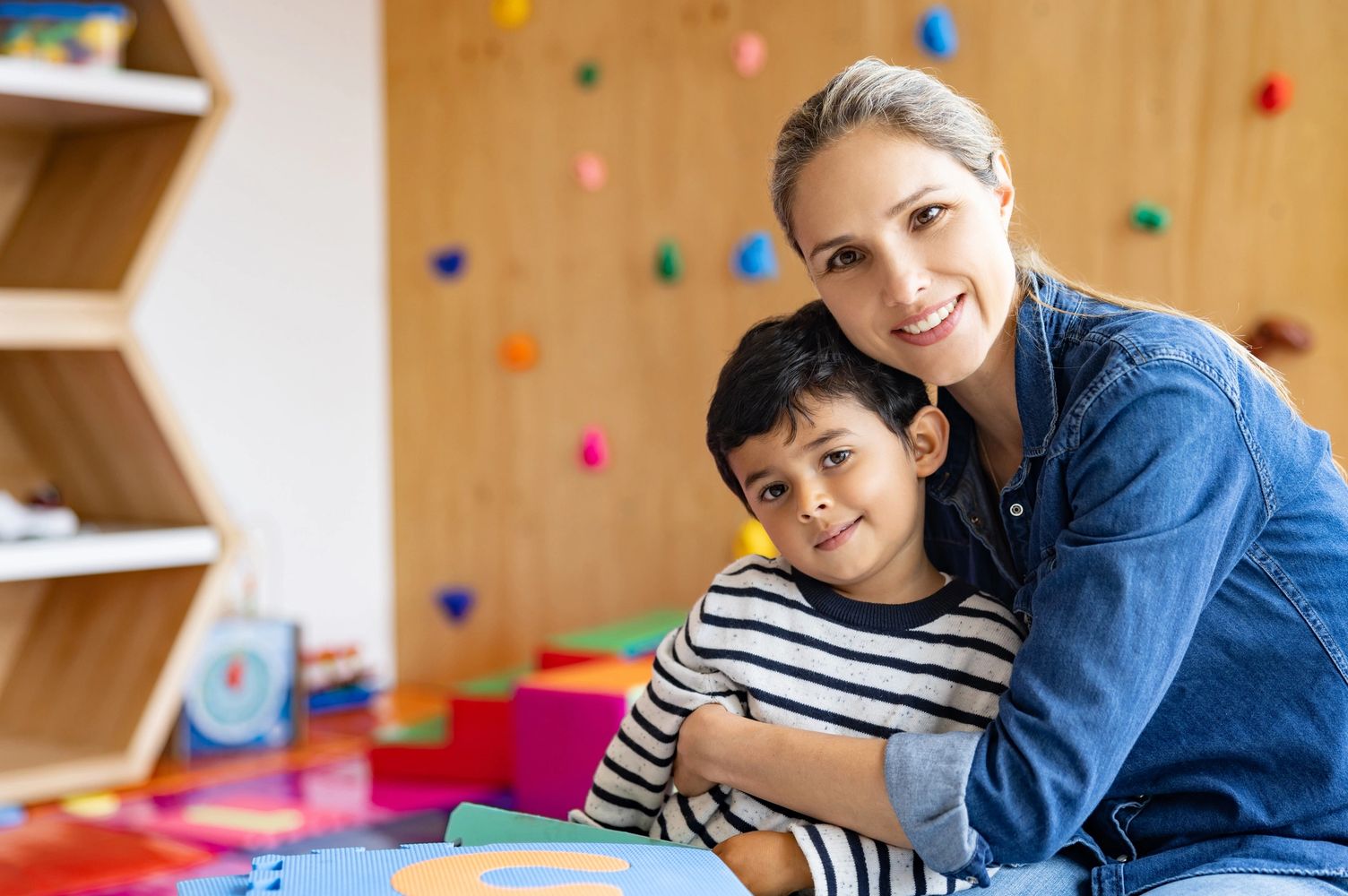 Woman and child smiling and hugging in a colorful playroom.